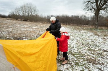 Bir adam ve genç bir kız, serin bir kış öğleden sonra çıplak ağaçlarla çevrili, karlı bir tarlada parlak sarı bir çadır kurmak için birlikte çalışırlar, neşe ve takım çalışmasını paylaşırlar..