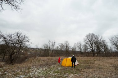 İki kişi, gri bir gökyüzünün altında, ferah, sessiz bir arazide, serin bir günde açık hava keşiflerinin ruhunu yakalayan, parlak sarı bir çadır kurdular..