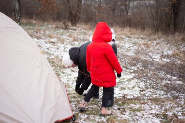 Serin bir kış öğleden sonrası, kıraç ağaçlarla çevrili, açık havada bir çadırın güvenliğini sağlayan parlak kırmızı paltolu bir çocuk olarak gözler önüne seriliyor..