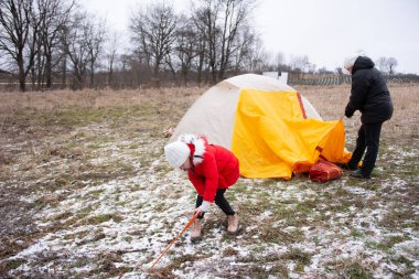 Çocuklar, çıplak ağaçlarla çevrili iken kışın serinliğini kucaklayan, karlı bir tarlada canlı bir çadır kurarak neşeli bir açık hava deneyimi yaratıyorlar..