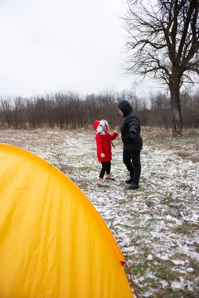 Parlak kırmızı bir palto giymiş bir çocuk, çıplak ağaçlar ve serin kış atmosferiyle çevrili, sarı bir çadır ile bir yetişkinle şakalaşarak etkileşime geçer..