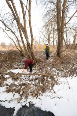 İki kişi nehir kenarını dikkatle temizlerken, bir diğeri de incelenen ağaçlar, karlı zemin ve kışın soğuğuyla ilgili ipuçlarıyla çevrili bölgeyi araştırıyor..