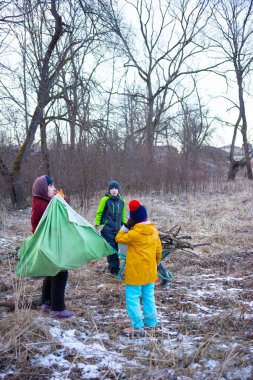 Bir grup çocuk, ilkbaharın başlarında çıplak ağaçlarla çevrili sakin, çimenli bir alanda çadır kurmalarına yardımcı olmak için dışarıda aktivitelere hevesle girişiyor..