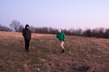 Alacakaranlıkta, iki arkadaş nazik bir yamaçta gülüşürler. Biri yeşil bir ceketle poz verirken, diğeri hareketsiz duruyor, doğanın sessiz güzelliğinde samimi bir karşılaşmayı somutlaştırıyor..