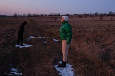 İki figürlü sakin bir kırsal manzara alacakaranlıkta gözler önüne seriliyor. Biri parlak yeşil bir ceketin içinde güvenle dururken, diğeri sönen ışığın ortasında düşüncelere dalmış, hafifçe eğilmiş..