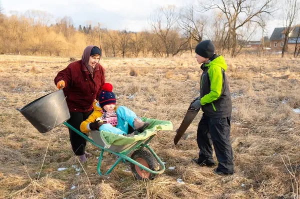 Güneşli bir günde çimen ve ağaçlarla dolu bir kırsal bölgede çocukların bir yetişkin tarafından el arabasına itilmenin heyecanını yaşadıkları neşeli bir ortam..