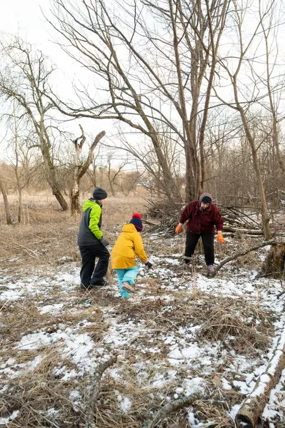 Çocuklar ve bir yetişkin, ağaçlar ve kuru otlarla çevrili, karlı vahşi doğada oynarken çubuk topluyor, doğanın kucaklamasında hoş bir kış keşfi yapıyorlar..