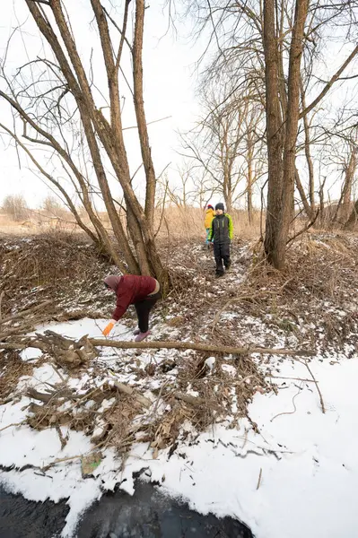 İki kişi nehir kenarını dikkatle temizlerken, bir diğeri de incelenen ağaçlar, karlı zemin ve kışın soğuğuyla ilgili ipuçlarıyla çevrili bölgeyi araştırıyor..