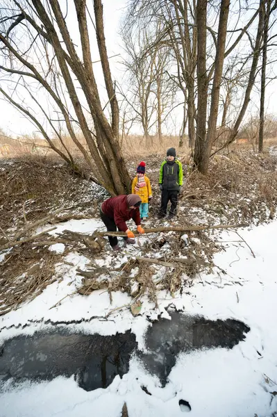 Üç birey kısmen donmuş bir derenin kenarında kış macerasının tadını çıkarırken, bir yetişkin kar ve dalları kazıyor ve çocuklar da merakla soğuğu seyrediyor..