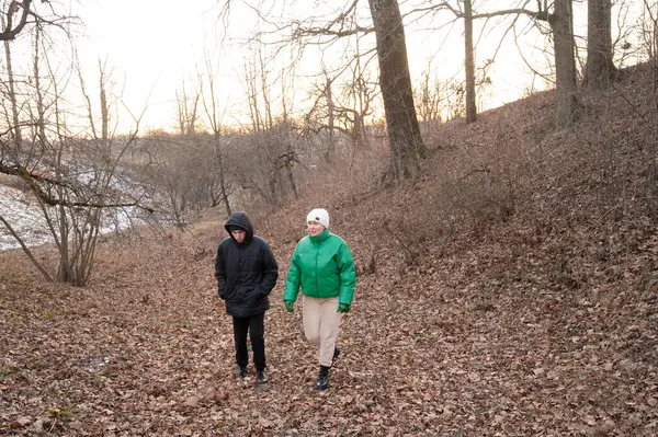 Bir çift arkadaş, kış ağaçlarıyla çevrili, sessiz, yapraklı bir patikada geziniyor. Günbatımının ılık parıltısı serin havayla çelişiyor ve sakin bir atmosfer yaratıyor..