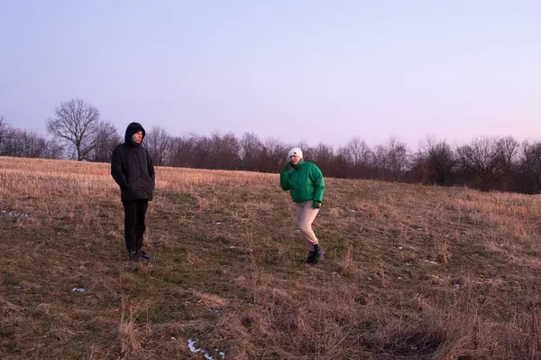 Alacakaranlıkta, iki arkadaş nazik bir yamaçta gülüşürler. Biri yeşil bir ceketle poz verirken, diğeri hareketsiz duruyor, doğanın sessiz güzelliğinde samimi bir karşılaşmayı somutlaştırıyor..