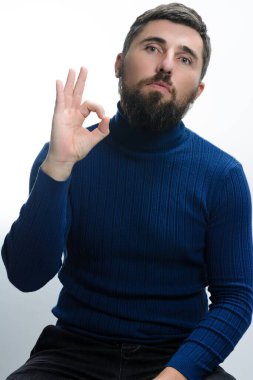 Mavi balıkçı yaka kazak ve koyu renk pantolon giyen sakallı bir adam elini açık arkaplana doğru uzatarak iyi bir jest yapıyor..