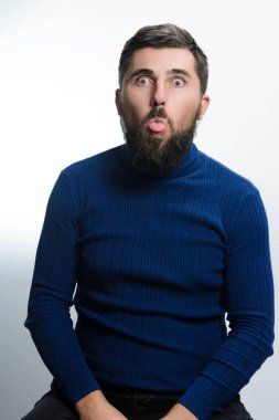 Mavi boğazlı kazaklı sakallı bir adam dilini beyaz arka plana doğru uzatıyor. Onun neşeli ifadesi mizah ve hafiflik taşır..