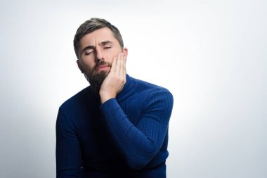 Mavi kazaklı sakallı bir adam, basit bir fon karşısında yanağını, gözlerini kapalı tutar. Yüzündeki ifade rahatsızlık ya da diş ağrısı olduğunu gösteriyor..