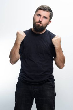 Siyah gömlekli ve siyah pantolonlu sakallı bir adam yumruklarını sıkarak hafif bir arka plana karşı kararlılık ya da saldırganlık hissini iletir..