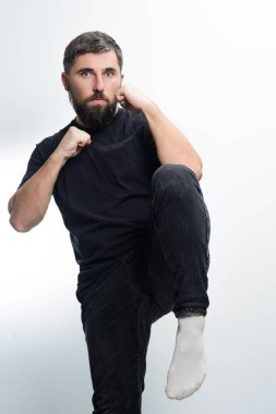 Siyah gömlek ve koyu renk kot giyen sakallı bir adam, düz beyaz bir arka plana karşı savaş duruşu takınır. Bir bacağını kaldırdı ve yumruklarını kaldırdı..