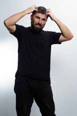 Siyah tişörtlü ve siyah pantolonlu sakallı bir adam açık gri bir arka plana karşı duruyor. Kafasını kaşıdığında açıkça hayal kırıklığına uğruyor..