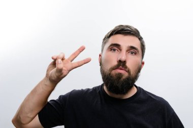 Parmaklarıyla barış işareti yapan sakallı bir adamın portresi. Siyah bir tişört giyer ve kameraya düz beyaz bir arka plana bakar..