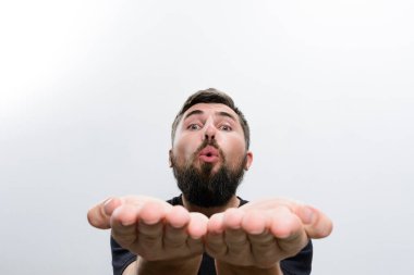 Beyaz arka planda sakallı bir adamın yakın plan fotoğrafı. Elleri birbirine kenetlenmiş bir şekilde kameraya öpücük yolluyor..