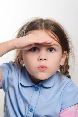 Kahverengi saçlı, açık mavi tişörtlü, meraklı bir çocuk, uzaktan bakıyor, eli gözlerini gölgeliyor. İfadesi konsantrasyon ve beklenti ile ilgili..