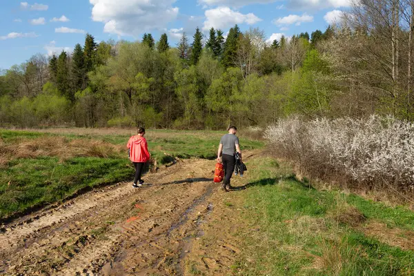 Bir çift arkadaş canlı yeşil bir arazide çamurlu bir yolda yürüyor, ağaçlar ve çiçek açan çalılarla birlikte baharın güzelliğinin tadını çıkarıyorlar..