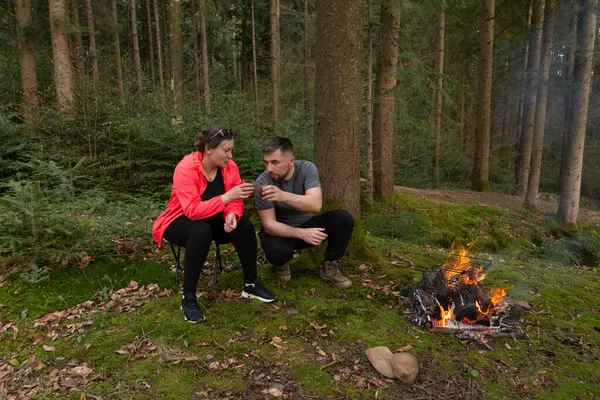 İki arkadaş yan yana oturur, içkilerini yudumlar ve alacakaranlıkta yükselen ağaçlar ve yemyeşil ağaçlarla çevrili bir kamp ateşinin sıcaklığının tadını çıkarırlar..