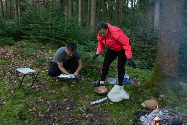 İki birey, sakin bir orman ortamında uzun ağaçlarla çevriliyken dışarıda yemek pişiriyor. Ateşin sıcaklığından ve birlikte yemek hazırlamanın heyecanından keyif alıyorlar..