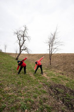 Canlı kırmızı giyinmiş iki kişi, seyrek yapraksız ağaçlarla çevrili sisli bir gökyüzünün altında çimenli bir yamaçta kollarını uzatıyorlar..