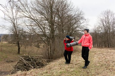 İki birey, serin ve bulutlu bir günde, doğanın güzelliğinin ortasında dinlendirici bir oturum için yoga minderleri taşıyarak sakin bir alanda yürürler..