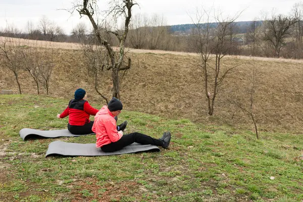 İki birey, baharın başlarında, seyrek ağaçlar ve nazik tepelerle çevrili, paspaslar üzerinde serinletici bir yoga seansının keyfini çıkarıyorlar..