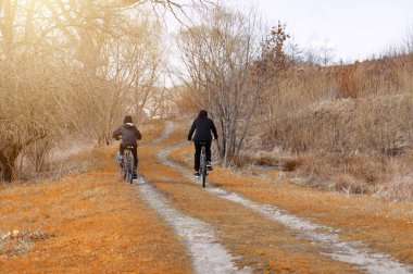 Bisikletlerdeki iki figür toprak bir yolda ilerliyor. Yol, sonbaharın veya kışın sonunun göstergesi olarak kurumuş, turuncu-kahverengi otlar ve yapraksız ağaçların arasından esiyor..