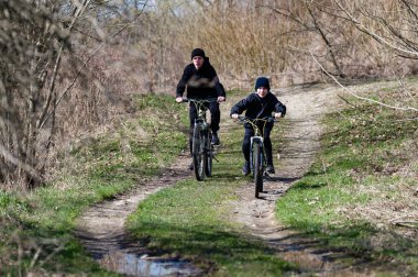 Tomurcuklanan bahar bitkilerinin arasında iki genç çocuk toprak bir patika boyunca bisiklet sürüyor. Güneş ışığı ağaçların arasından akar, parlak ve neşeli bir atmosfer yaratır. Dağ bisikleti sürüyorlar..