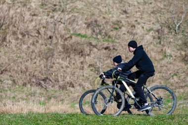 Çimenli bir yamaç boyunca iki kişi bisiklet sürüyor. Arka plan kuru, kahverengi bitkileri gösteriyor. Sahne bir özgürlük ve açık hava aktivitesi hissi veriyor..