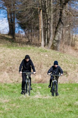 İki genç insan çimenli bir yolda bisiklet sürüyor, gün ışığında banyo yapıyorlar. Ağaçlar bisiklet sürüşleri için bir zemin oluştururlar. Kaygısız, aktif bir sahne..