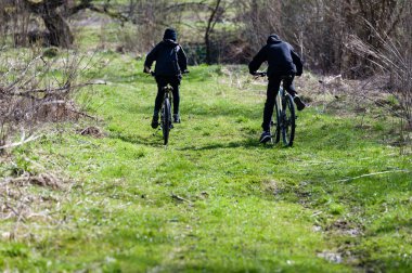 Çimenli bir yolda bisiklet süren iki kişinin dikiz görüntüsü. Güneş ışığı manzarayı aydınlatarak doğal çevrede barışçıl bir atmosfer yaratır. Bisikletçiler koyu renk giyiyorlar..