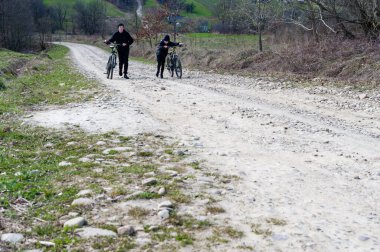 Bisikletlerini kırsal, çakıllı bir yolda yürüten iki kişinin manzarası. Mekan barışçıl ve kırsal, kırsal bir ortam olduğunu gösteriyor..