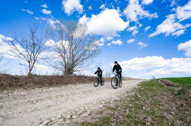 Canlı mavi bir gökyüzünün altında taşra çakıl yolunda bisiklet süren iki kişinin manzarası. Manzara sakin, ağaçlar ve tarlalar seyrek..