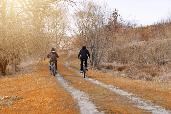 Bisikletlerdeki iki figür toprak bir yolda ilerliyor. Yol, sonbaharın veya kışın sonunun göstergesi olarak kurumuş, turuncu-kahverengi otlar ve yapraksız ağaçların arasından esiyor..