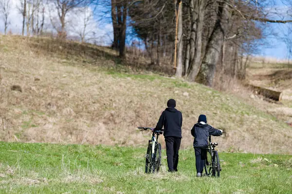 İki birey, muhtemelen baba ve oğul otlak bir yolda yürüyorlar. Bisikletler yedekte. Huzurlu kırsal alan, güneş ışığıyla yıkanmış. Tepeler ve ağaçlar etraflarını sarmış.