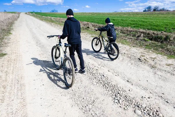 Bisikletleriyle çakıllı bir yolda yürüyen iki genç insan. Ortam kırsal, arka planda yeşil alanlar ve açık mavi bir gökyüzü var. Güneşli bir gün.