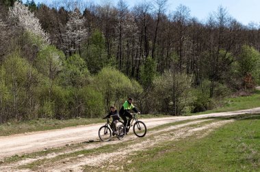 Bir adam ve çocuk, ağaçlar ve yeşilliklerle çevrili kırsal bir yolda bisiklet sürüyor. Güneş ışığı yaprakların arasından akar ve barışçıl bir atmosfer yaratır..