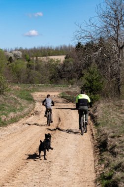 İki bisikletçi ve köpekleri, ormanlık alanda çamurlu bir yolda güneşli bir öğleden sonra bisiklet sürüşünün keyfini çıkarıyorlar. Ortam huzurlu ve neşeli..