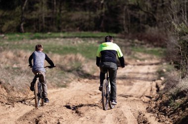 İki bisikletçi, muhtemelen bir baba ve oğul, kumlu bir yolda bisiklet sürüyorlar. İzler, arka planda ağaçlar ve bitki örtüsü olan doğal bir ortamda esiyor. Güneş ışığı sahneyi aydınlatır..