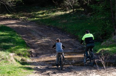 Bisikletlerdeki iki figür ormanlık alanda çamurlu bir patikadan geçiyor. Yaşlı birey, genç olanı yönlendirir ve paylaşılan açık hava aktivitelerinin iç açıcı bir görüntüsünü yaratır..