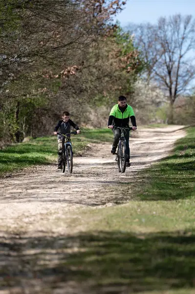 Bir adam ve bir çocuk, ağaçlar ve ilkbahar yapraklarıyla çevrili bir kır yolunda birlikte bisiklet sürüyorlar. Sahne, aile bağlarını ve açık hava aktivitelerini çağrıştırıyor..