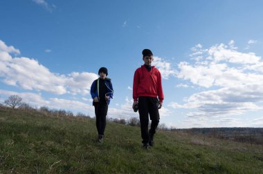 Sıcak giyimli iki genç, güneşli bir günde çimenli bir yamaçta yürüyorlar. Gökyüzü parlak bir mavi tüylü beyaz bulutlarla.