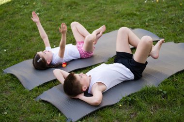 İki çocuk, bir erkek ve bir kız, çimenli bir alanda yoga minderleri üzerinde egzersiz yapıyorlar. Fitness rutininden zevk alıyor gibi görünüyorlar..