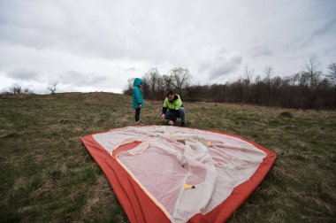 Bir kişi ve bir çocuk, bulutlu gökyüzü ve doğanın güzelliği ile çevrili çimenli bir alanda renkli bir çadır kurmak için birlikte çalışırlar. Takım çalışmasını ve macerayı teşvik ederler..