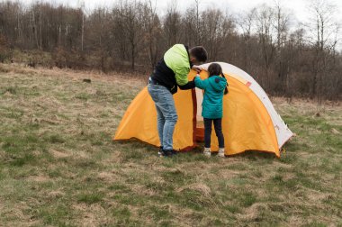 İki kişi parlak turuncu bir çadır kurmakla meşgul. Serin bir bahar gününde, çıplak ağaçlar ve çimenli tarlalarla çevrili sakin bir açık alan..