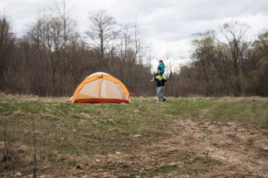 Bir çocuk taşıyan bir kişi bulutlu bir günde ağaçlar ve çimenlerle çevrili sakin doğal bir ortamda kurulmuş parlak turuncu bir çadıra doğru yürür..